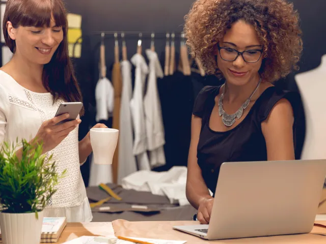 Deux femmes au comptoir d'un magasin de vêtements. L'une consulte son téléphone et l'autre son laptop au sujet de l'adaptation des statuts de société en France (obligation légale à réaliser avant le 1er janvier 2024)